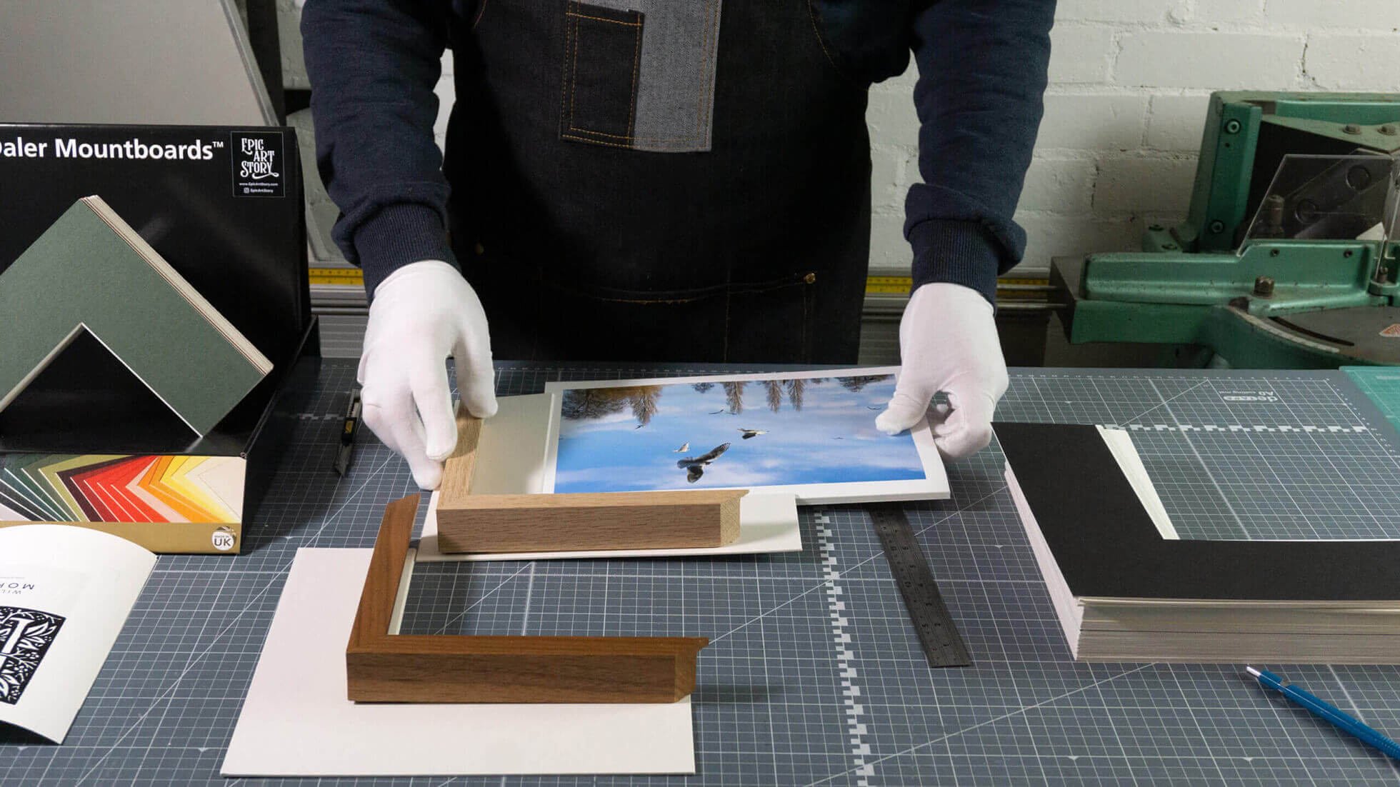 Professional framer wearing conservation gloves handling fine art photography with wooden frame samples at Epic Art Story workshop in Molesey